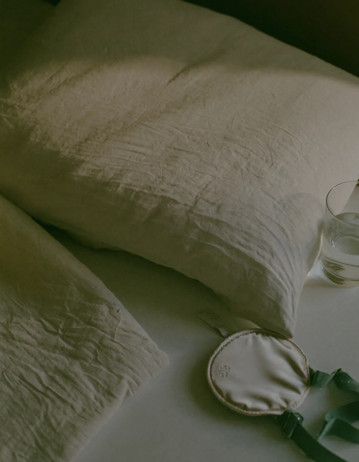 A rumpled linen pillowcase, a silk sleep mask and a glass of water on a bedside surface in dim early light