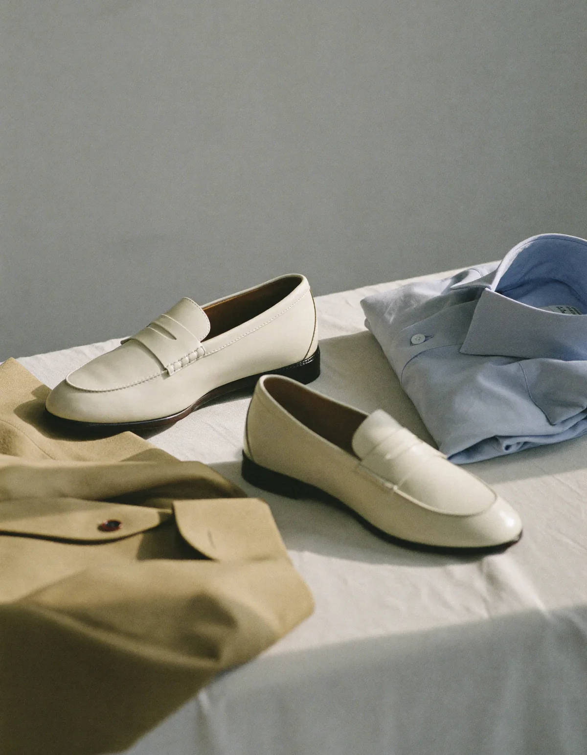 A folded camel blazer beside ivory leather loafers and a folded pale blue shirt on pale oyster linen