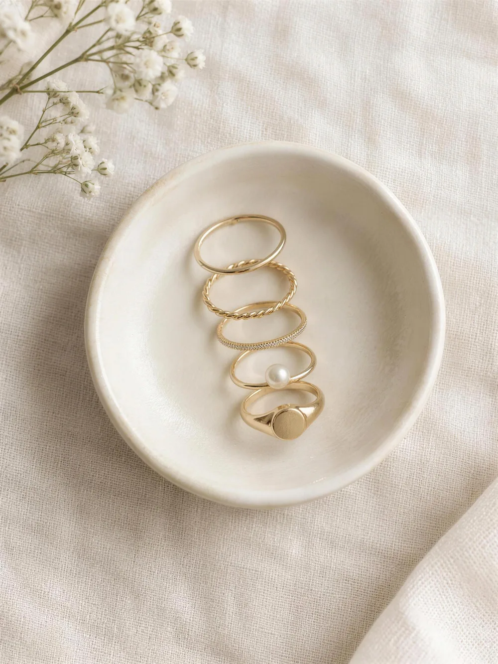 A line of delicate gold stacking rings in a cream ceramic dish: plain, twisted, pave, pearl and signet bands