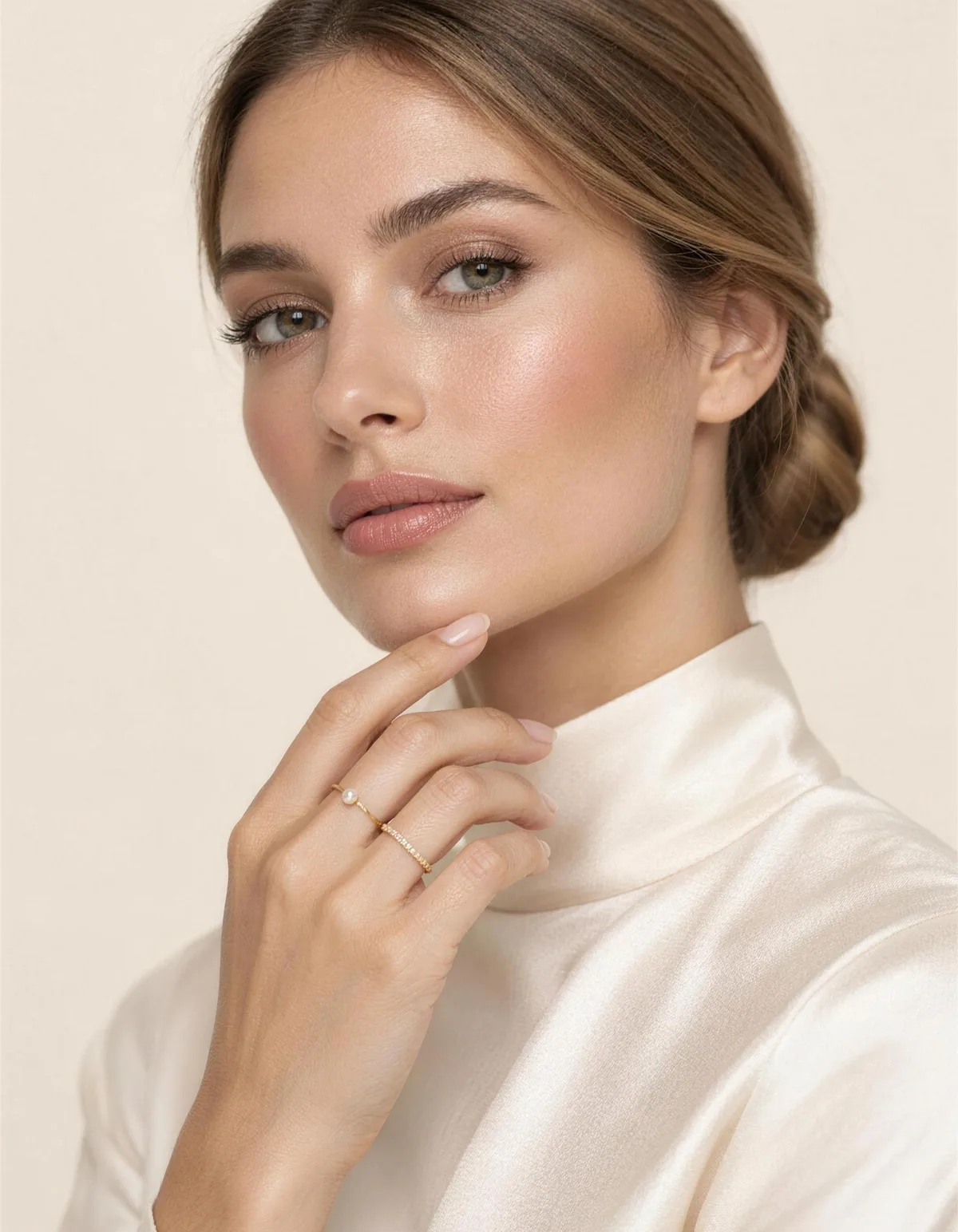A woman with a sleek low chignon touching her chin, wearing thin gold stacking rings with a tiny pearl and a pave band