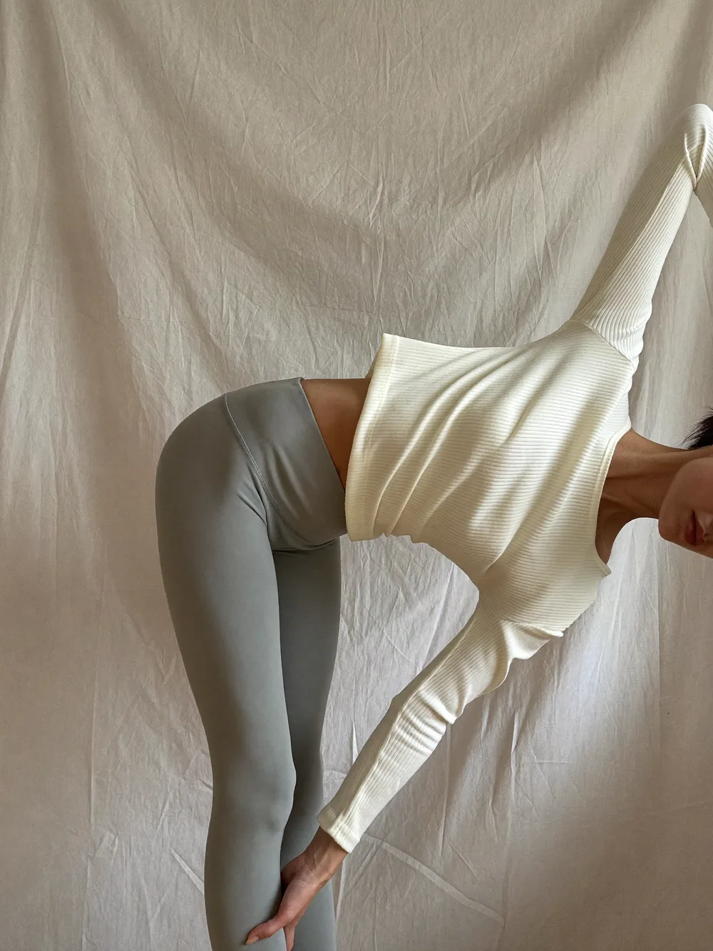 A woman in a cream long-sleeve top and sage leggings doing a standing side bend with one arm overhead