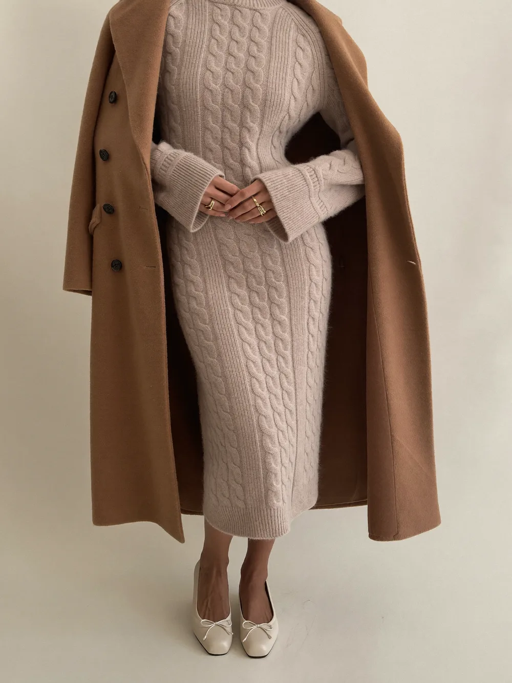 A woman in an oatmeal cable-knit midi sweater dress with a camel coat draped over her shoulders and cream flats