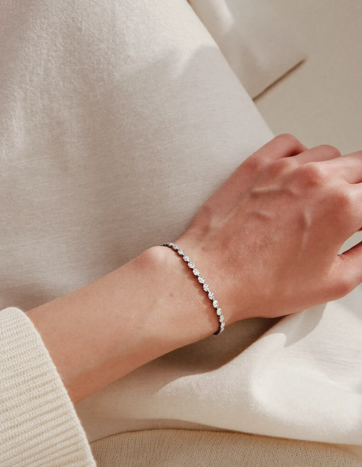 A slim wrist resting on cream linen wearing a delicate diamond tennis bracelet in white gold