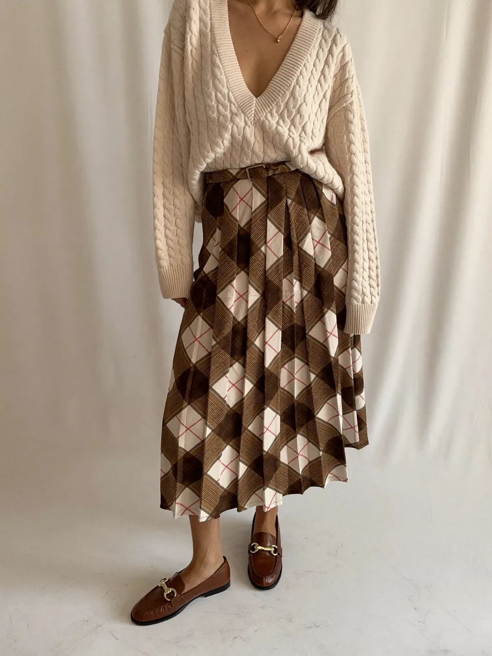 A woman in a cream cable-knit sweater tucked into a brown plaid pleated midi skirt with brown loafers