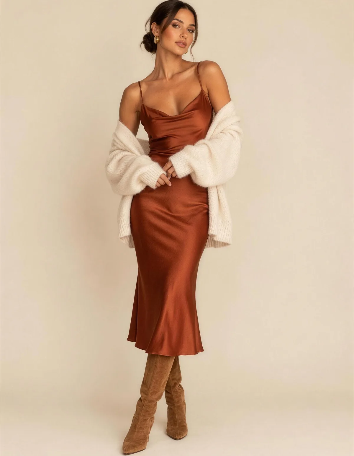 A beautiful woman in a rust satin slip midi dress with a cream cashmere cardigan draped off her shoulders and tan suede boots