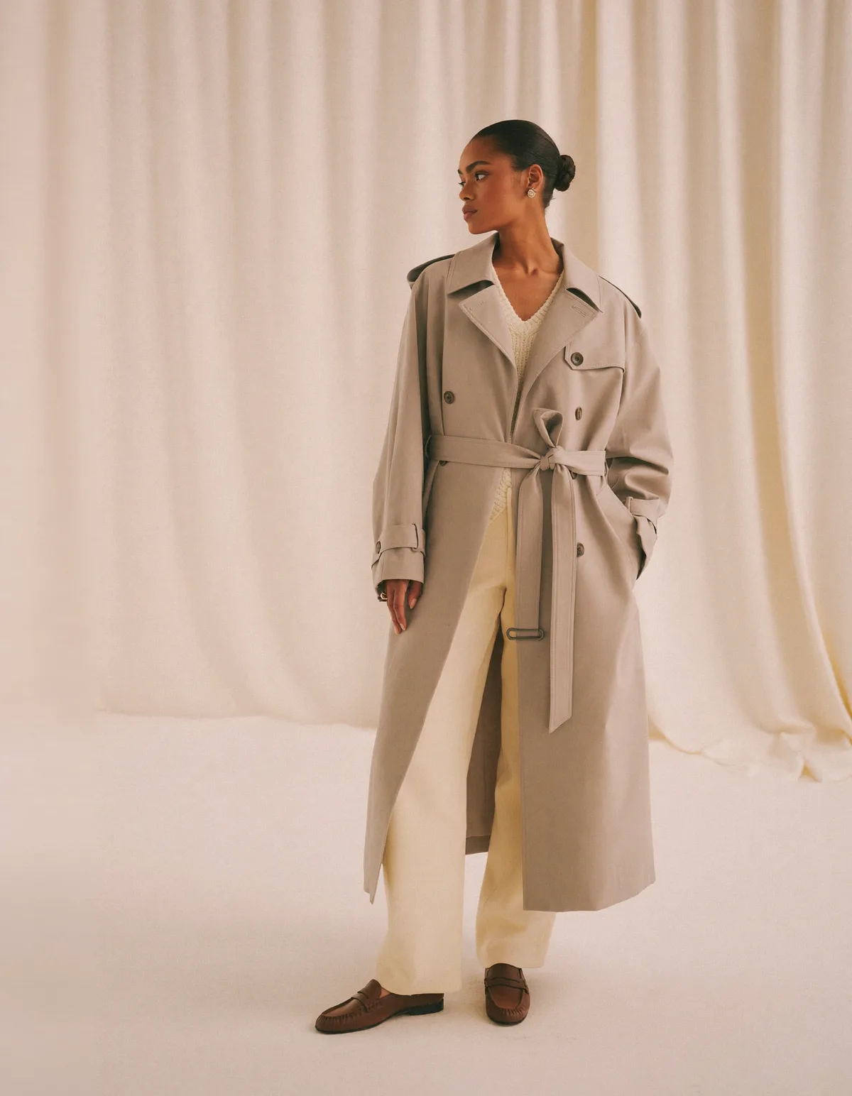 A woman with a sleek low bun in a belted beige trench coat over a cream knit and straight cream trousers