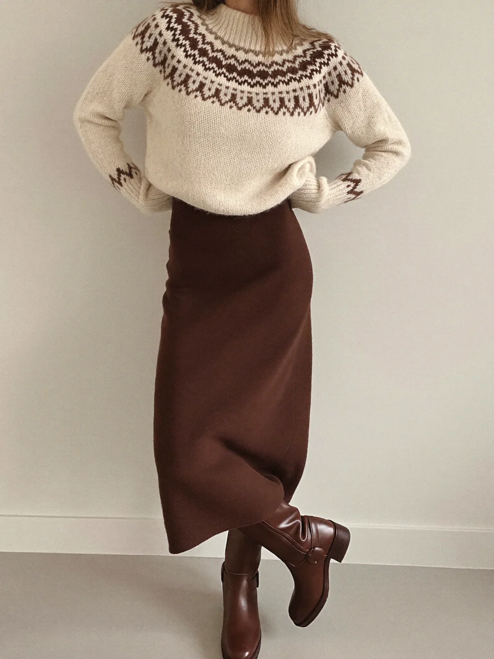 A woman against a cream wall in a cream and brown Fair Isle sweater, chocolate midi skirt and knee-high boots