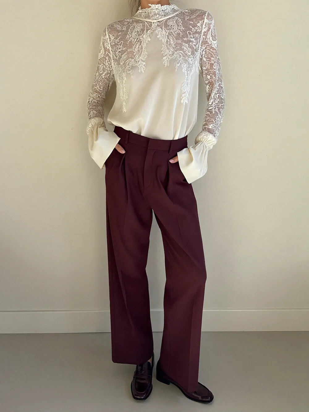 A woman against a cream wall in an ivory high-neck lace blouse tucked into burgundy wide trousers with burgundy loafers