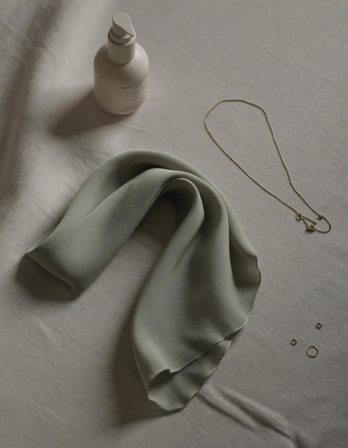 A folded sage silk scarf, a slim gold chain necklace and a small bottle of neck cream on pale oyster linen