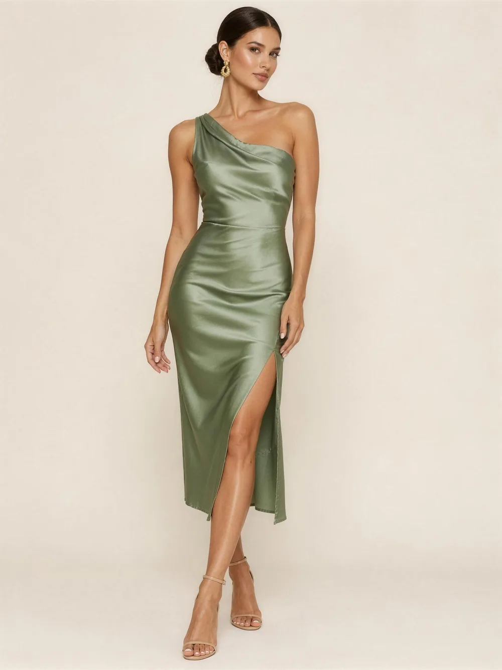 A woman in a figure-skimming sage green satin one-shoulder midi dress with a drape and a slit, gold earrings and nude heels