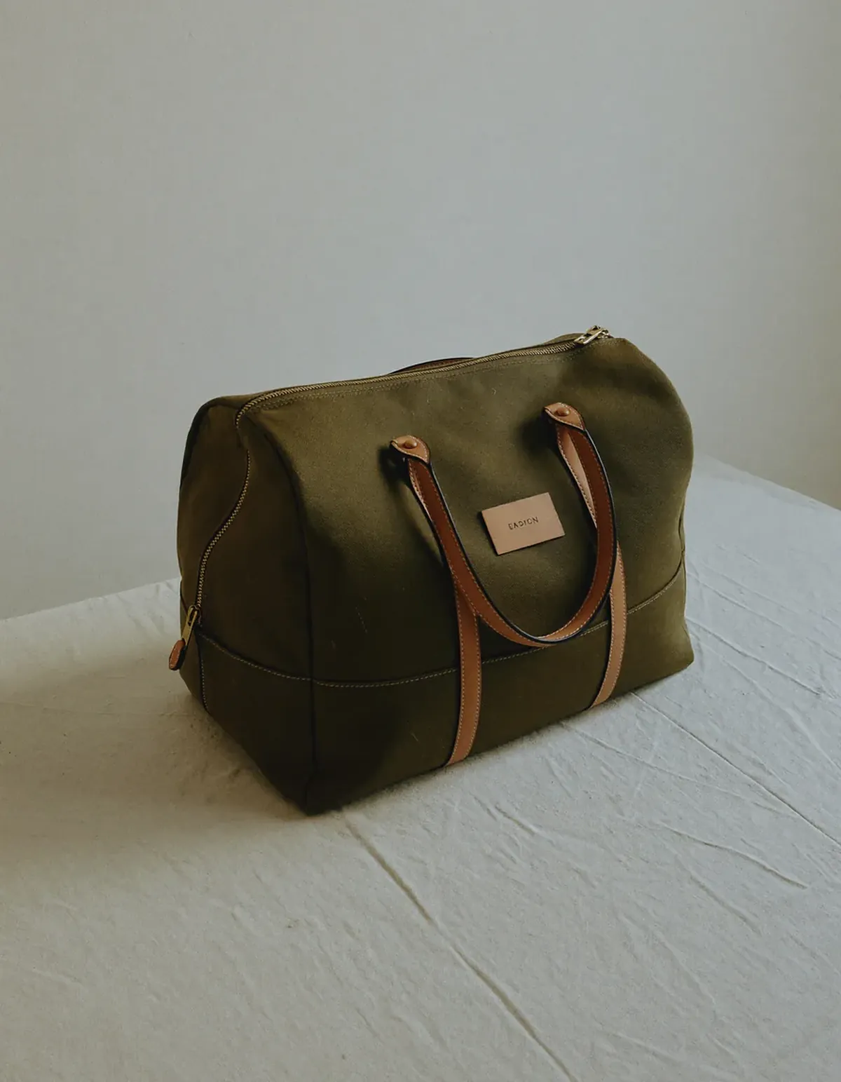 A waxed olive canvas weekender duffle bag with tan leather handles on pale oyster linen
