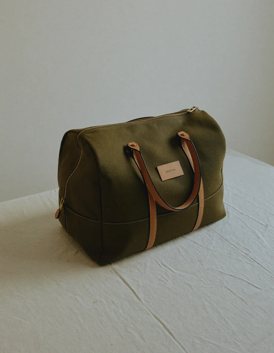 A waxed olive canvas weekender duffle bag with tan leather handles on pale oyster linen