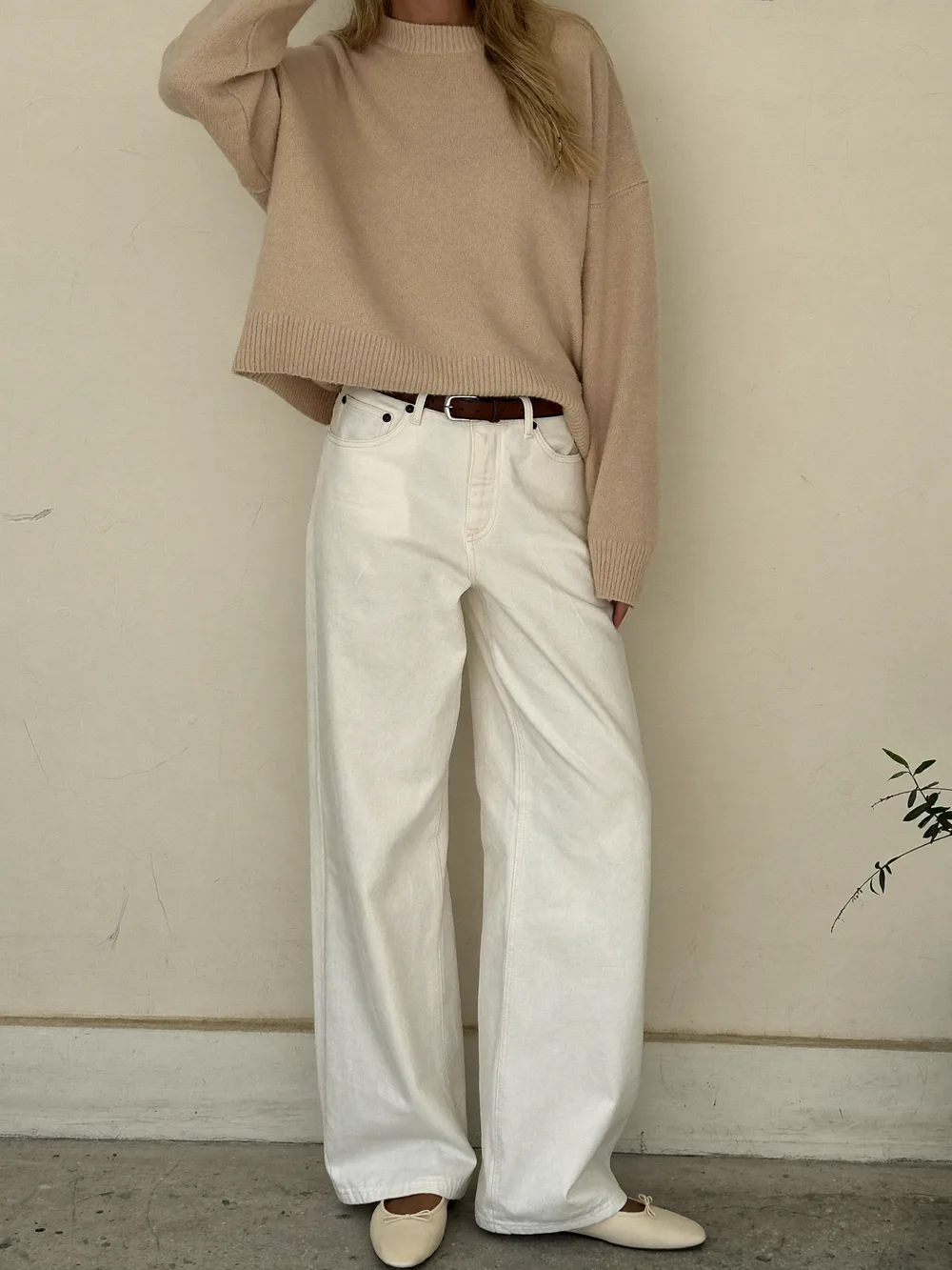 A woman in wide leg ecru jeans, a relaxed sand cashmere sweater with a brown belt and cream ballet flats against a cream wall