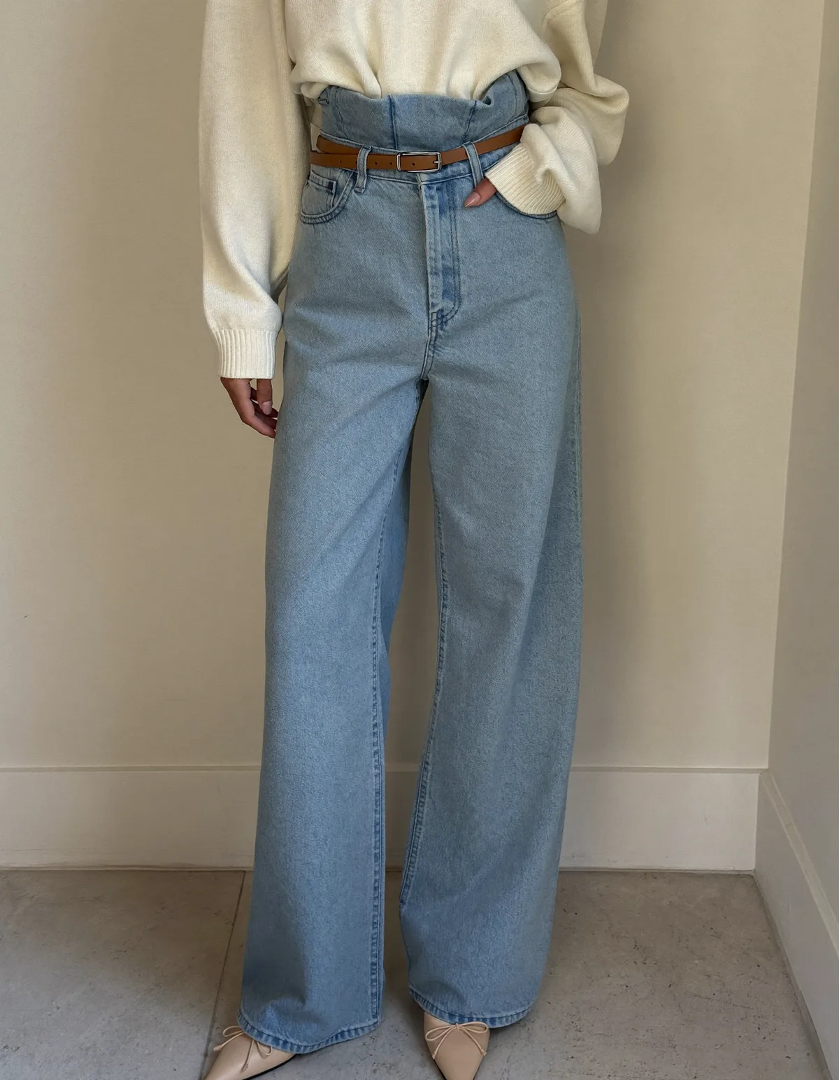 A woman against a cream wall in high-rise light blue wide leg jeans, a tucked cream knit, a tan belt and pointed flats