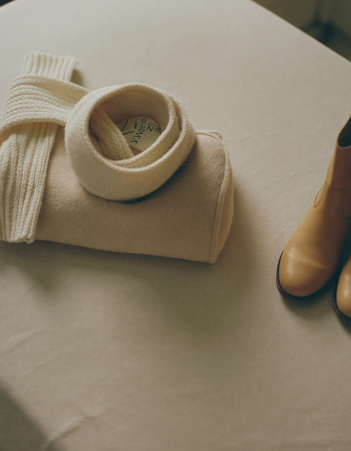 A folded oatmeal wool coat, a chunky cream knit, a wool scarf and tan leather boots on pale oyster linen